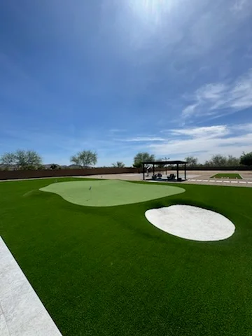 Synthetic golf putting green with sand bunkers and covered pavilion structure under blue sky