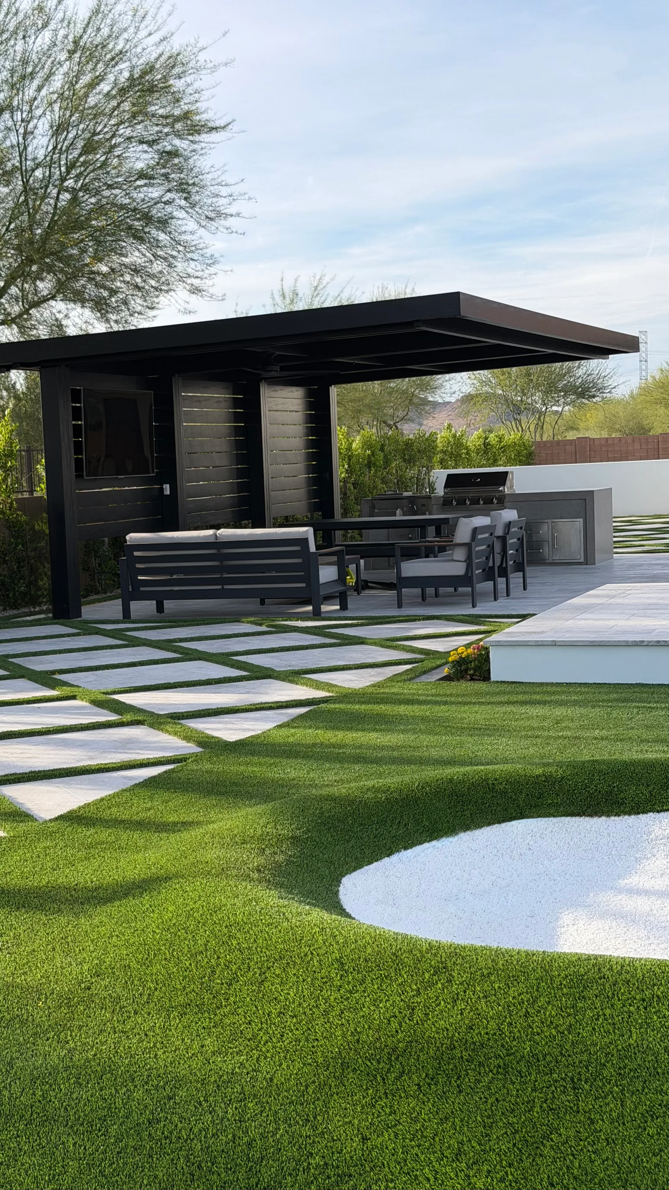 Modern black pergola with outdoor kitchen and geometric paver patio with grass strips